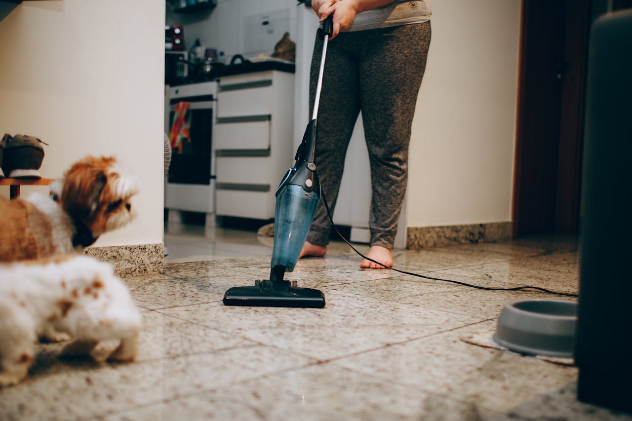 services-02 A woman cleaning the floor with a vacuum cleaner while a dog watches attentively. Indoor home setting.