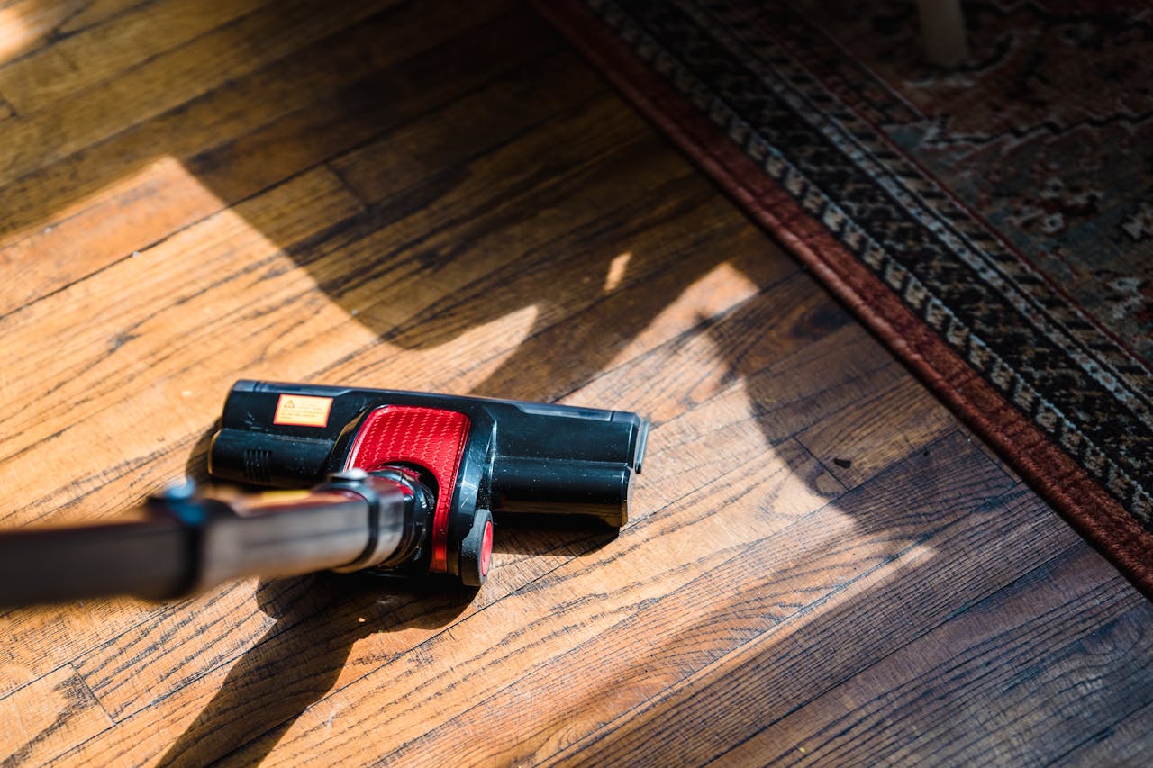 gallery-6 Close-up of a vacuum cleaner on wooden flooring near a patterned rug, showcasing domestic cleaning.